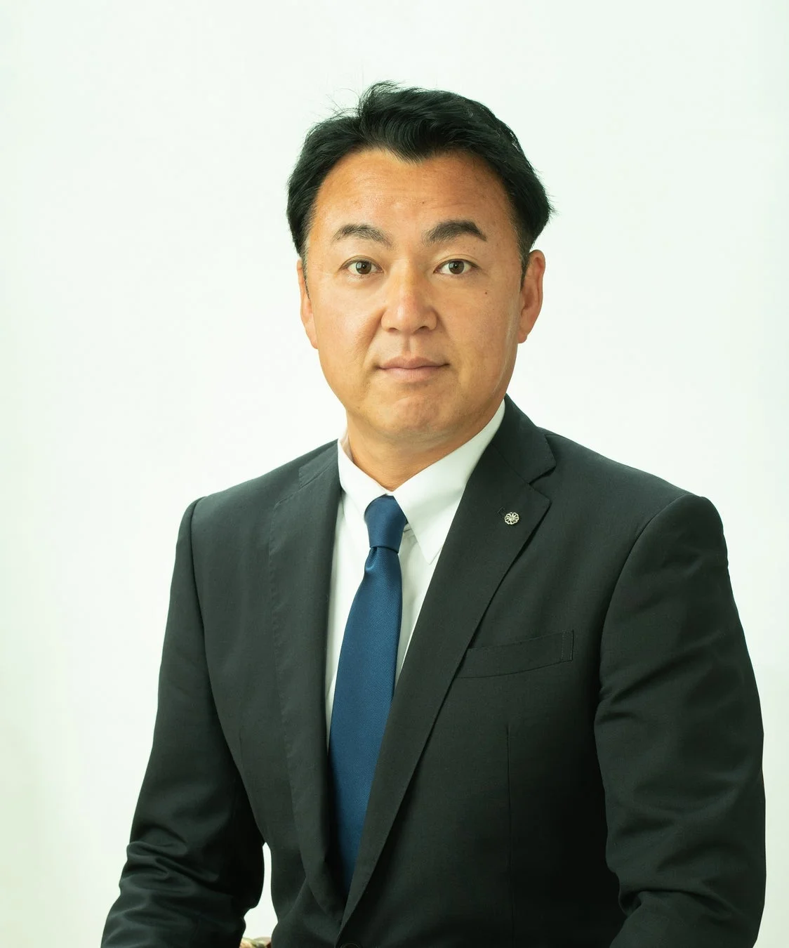 黒いスーツと青いネクタイを着用したアジア人男性が、白い背景の前でカメラを見つめている上半身のポートレート写真です。