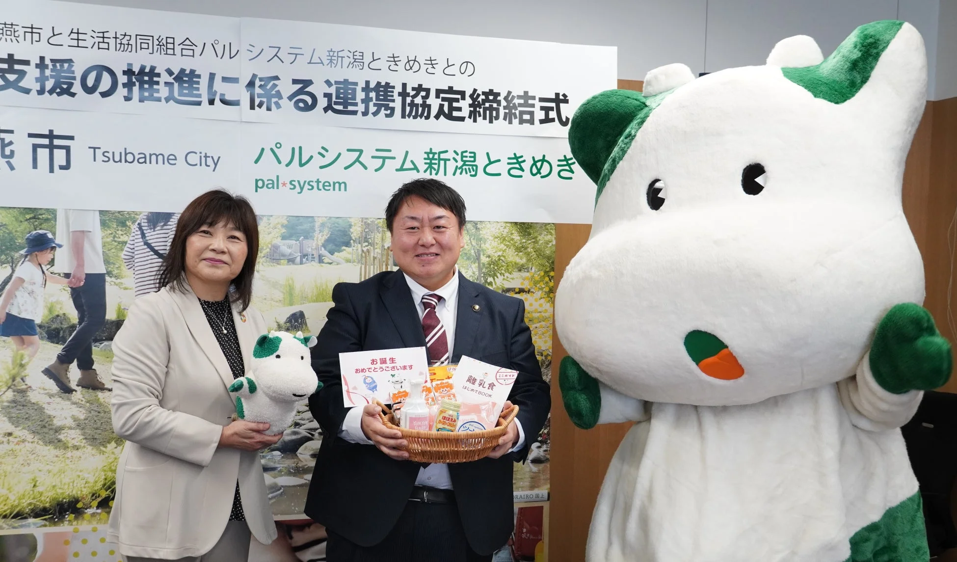 燕市と生活協同組合パルシステム新潟ときめきによる連携協定締結式の様子。男性と女性が笑顔で、マスコットキャラクターと共に記念撮影をしています。男性は赤ちゃん用品の入ったバスケットを手にしています。